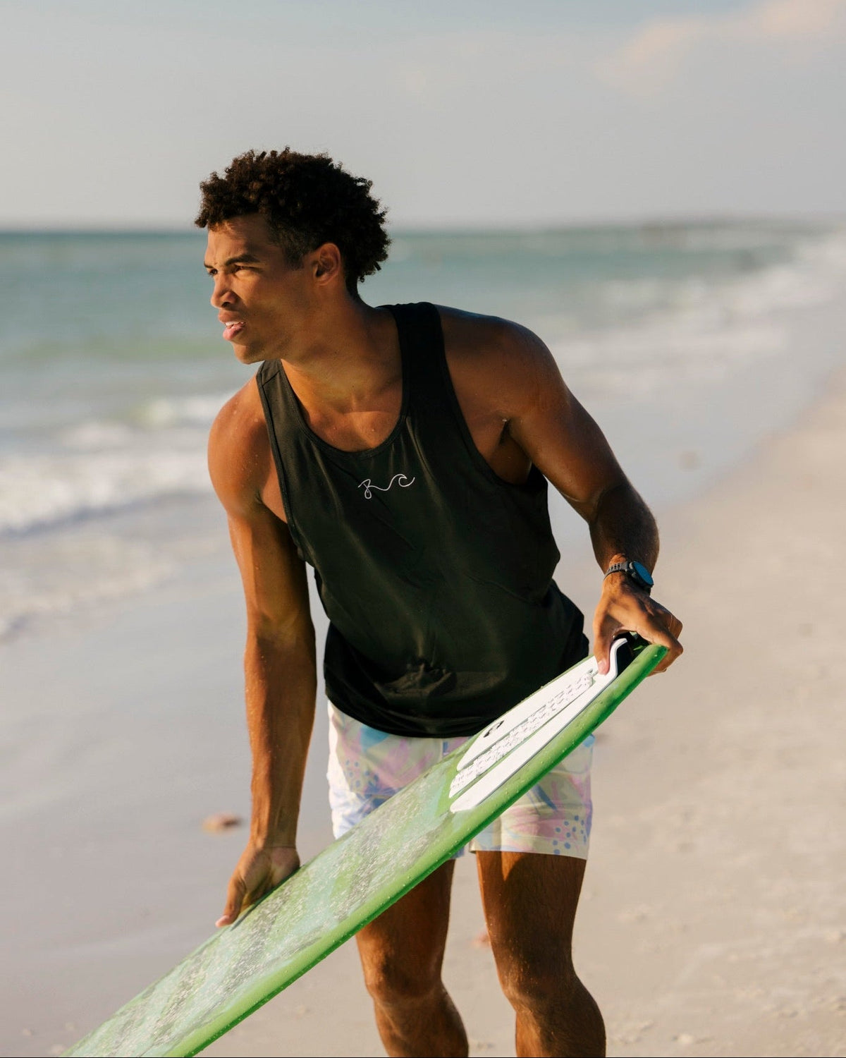 A man on a sandy beach walks toward the ocean holding a green surfboard. He is wearing a dark tank top and white patterned board shorts.