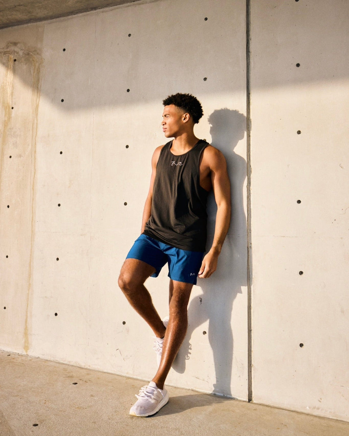 A man wearing a black athletic tank top and blue athletic shorts leans against a concrete wall in a sunlit outdoor setting.
