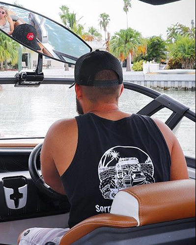 Two men wearing matching black tank tops with a white graphic and the text 'Sail For The Wake' on the back, sitting inside a boat. The driver wears a black cap and the passenger wears a blue cap.