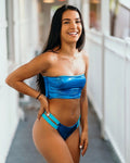 A woman modeling a shiny blue two-piece bikini with a bandeau-style top and high-cut bottoms with a light blue side accent, standing in a brightly lit hallway.