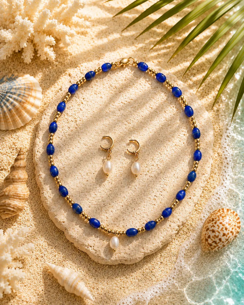 A gold and blue beaded necklace with matching drop earrings featuring white pearls, styled on a stone over a sandy beach background with seashells and palm leaves.