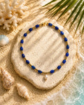 A beaded necklace with deep blue round beads, small gold spacer beads, and a single white pearl pendant, arranged on a stone over sandy beach decor with shells and palm leaves.