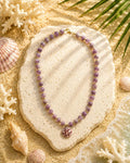 A beaded necklace with lavender-purple round beads and a small pink shell pendant, displayed on a textured stone slab over a sandy beach-style surface with seashells and palm leaves.