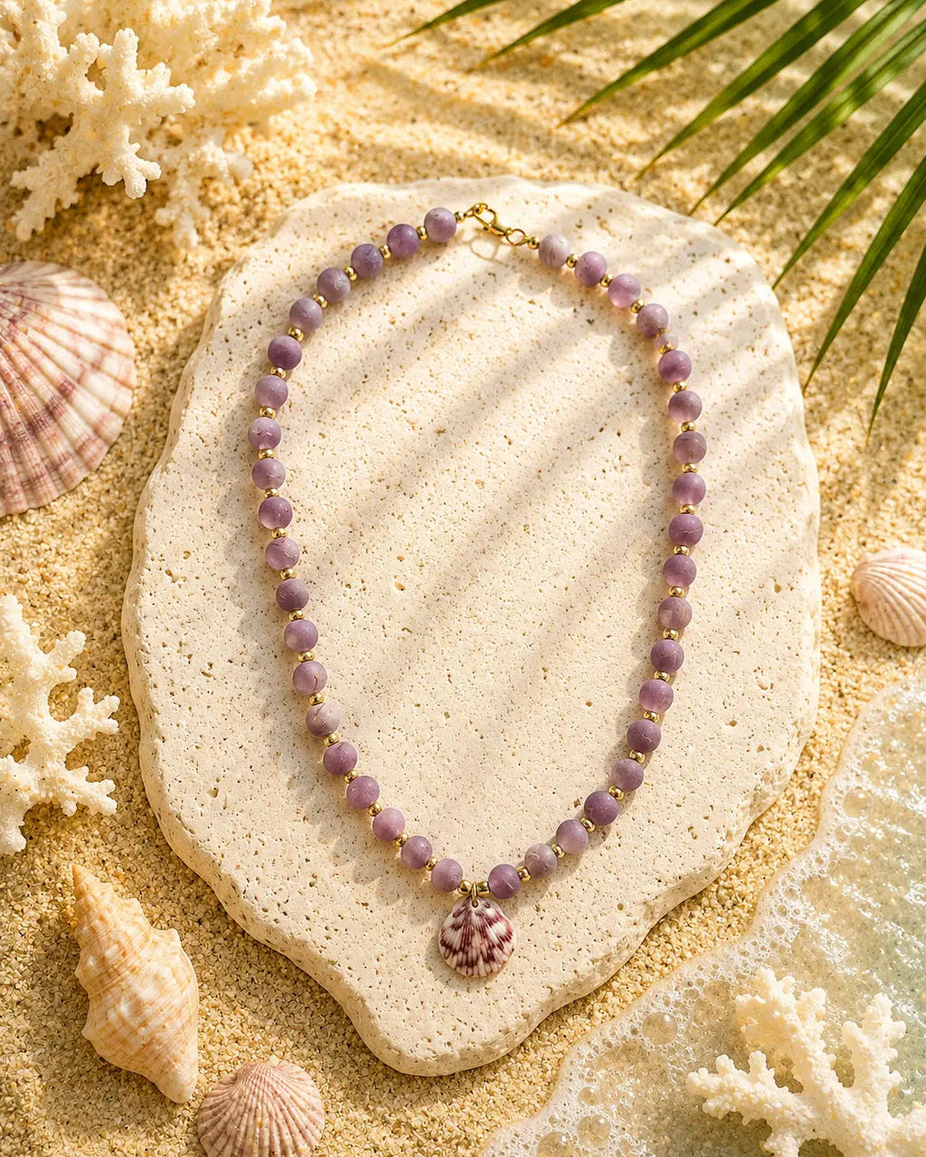 A beaded necklace with lavender-purple round beads and a small pink shell pendant, displayed on a textured stone slab over a sandy beach-style surface with seashells and palm leaves.