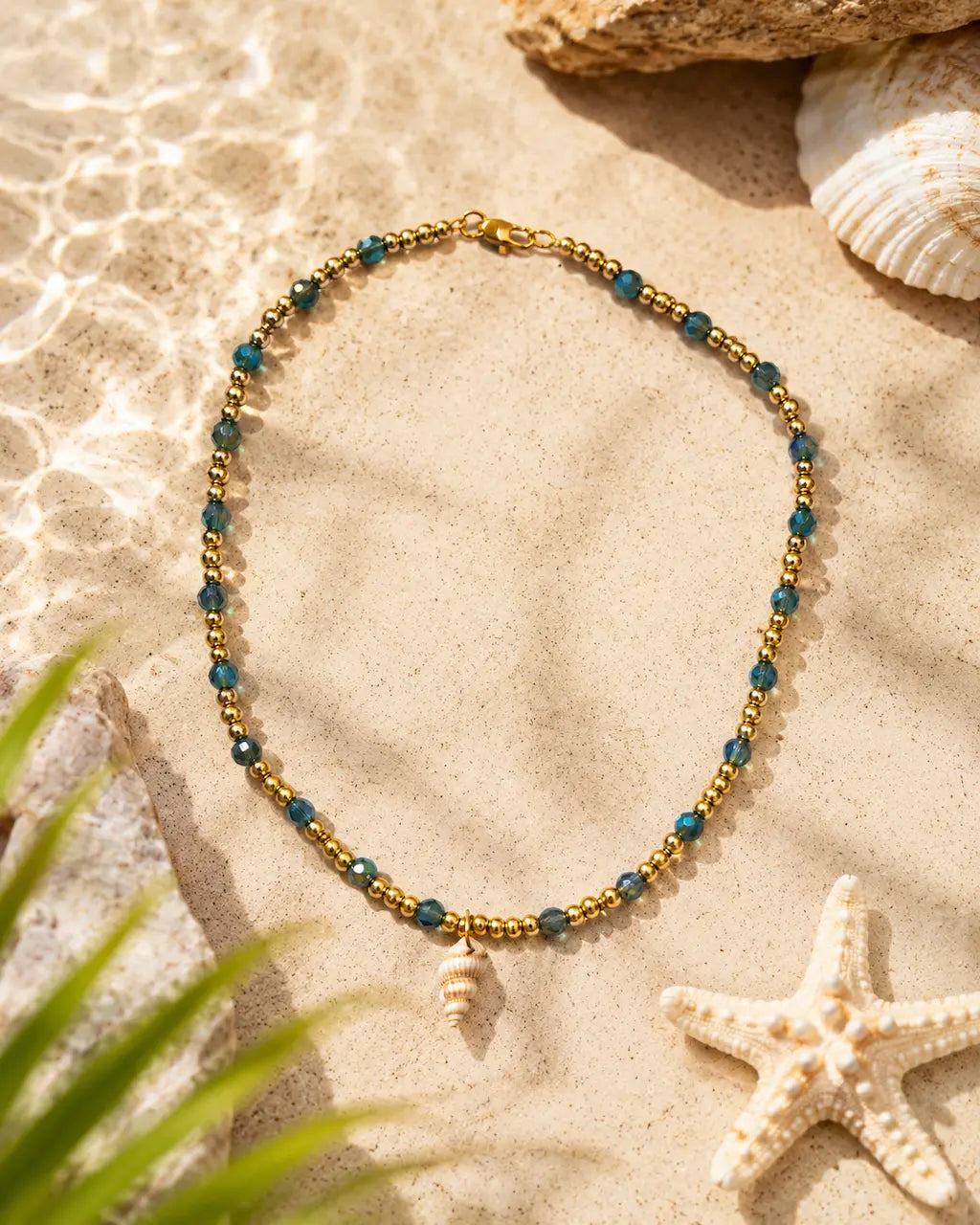 A gold and teal beaded necklace with a small seashell charm, styled on a sandy beach background with a starfish, seashells, and green leaves.
