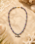 A beaded necklace with lavender purple round beads, iridescent blue-green faceted beads, and a small silver shark charm, displayed on a sandy surface with seashells and coral props.