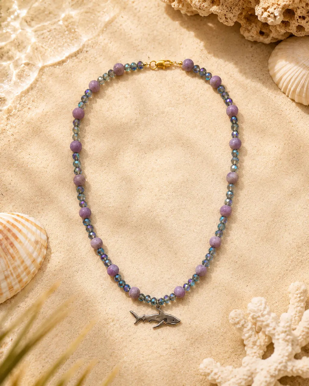 A beaded necklace with lavender purple round beads, iridescent blue-green faceted beads, and a small silver shark charm, displayed on a sandy surface with seashells and coral props.