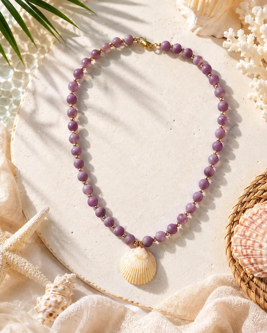 A beaded necklace with round lavender-purple beads and a seashell pendant, styled on a light beige surface with seashells, fabric, and green leaves.