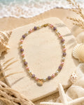 A delicate beaded necklace with lavender, pink, white, and gold beads, styled on a stone block with seashells and starfish against a beach background.