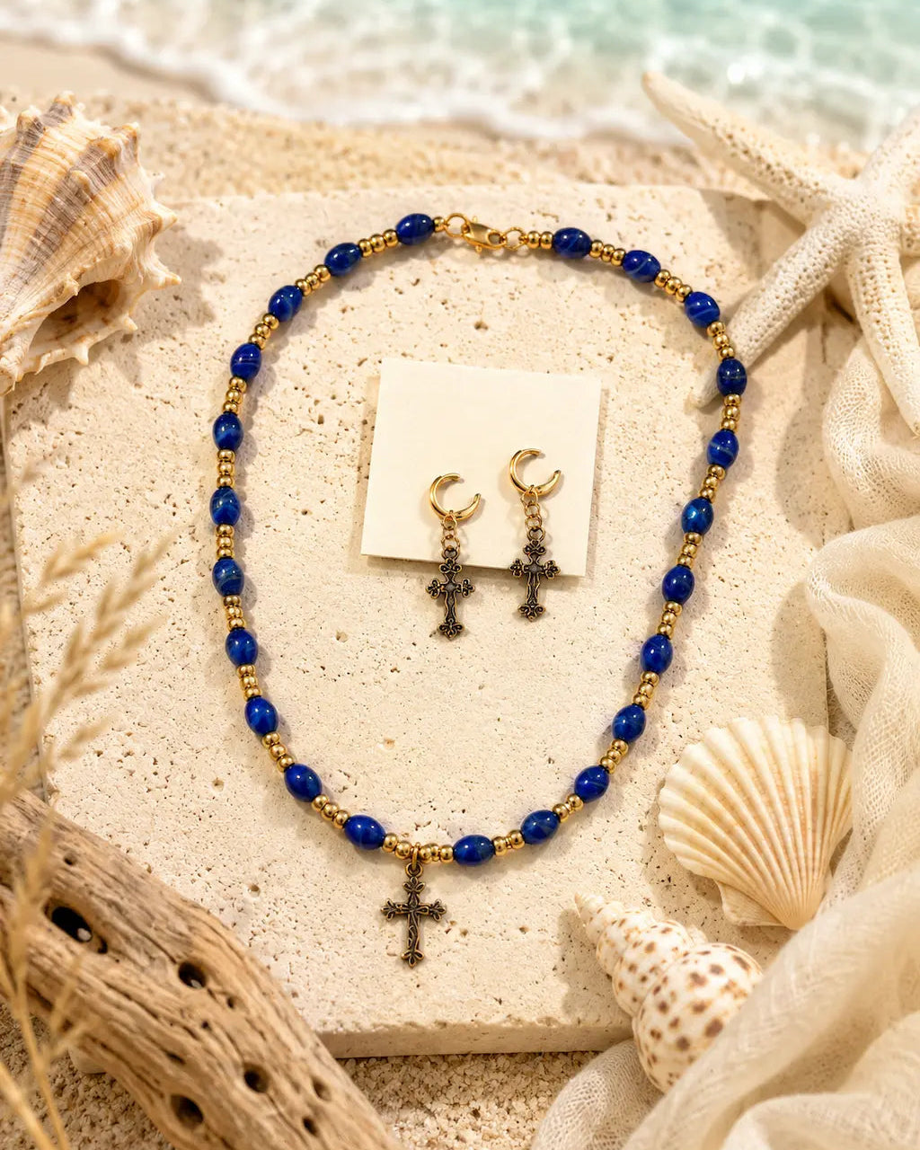 A beaded necklace and matching drop earrings displayed on a beige stone slab with seashells and fabric, featuring blue beads, gold spacer beads, and dark cross pendants.