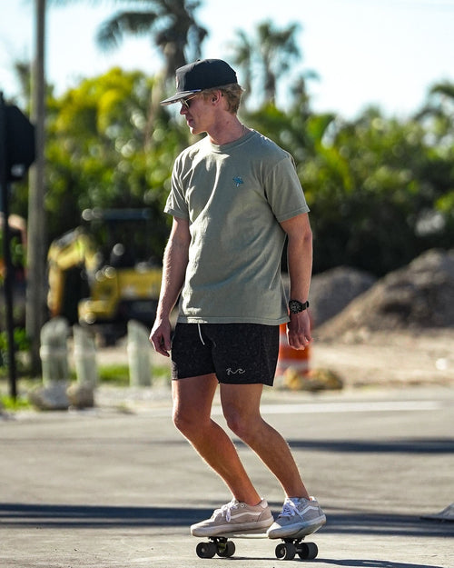 A man wearing a light olive green t-shirt, black shorts, and a dark cap is riding a skateboard on an outdoor paved road with palm trees in the background.