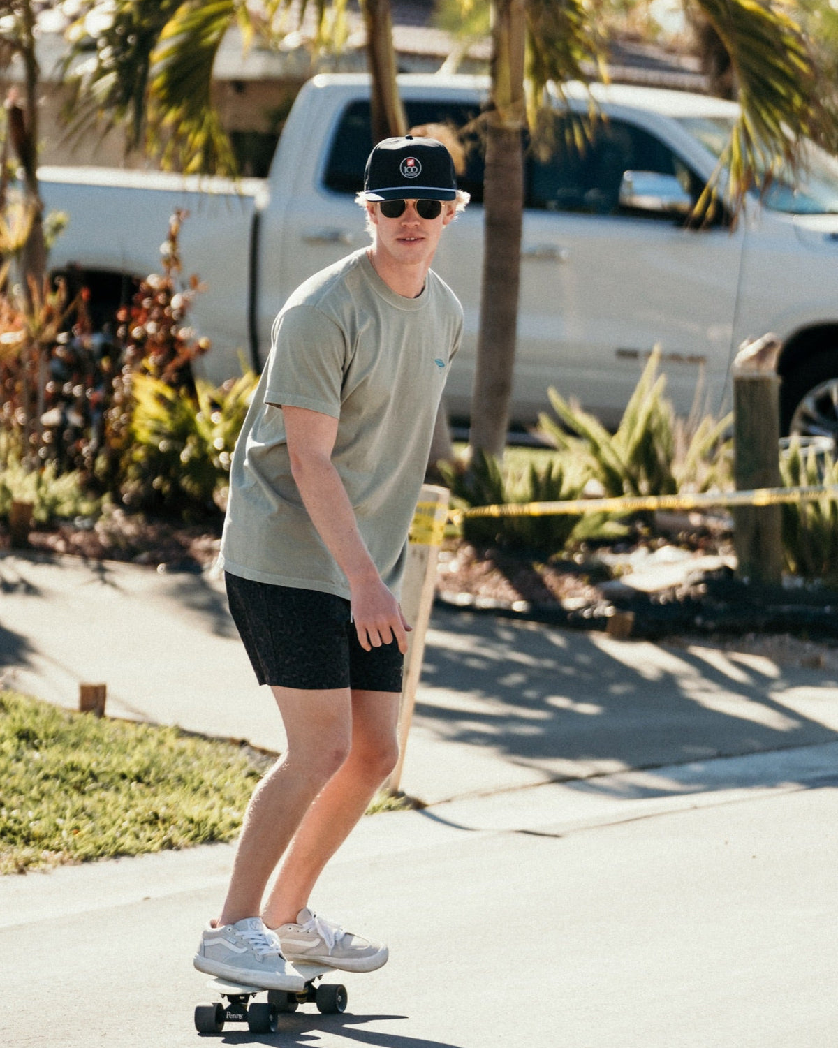 A man wearing a light olive green t-shirt, black shorts, white sneakers, and a black baseball cap rides a longboard skateboard on a sunny residential street with palm trees and a white pickup truck in the background.