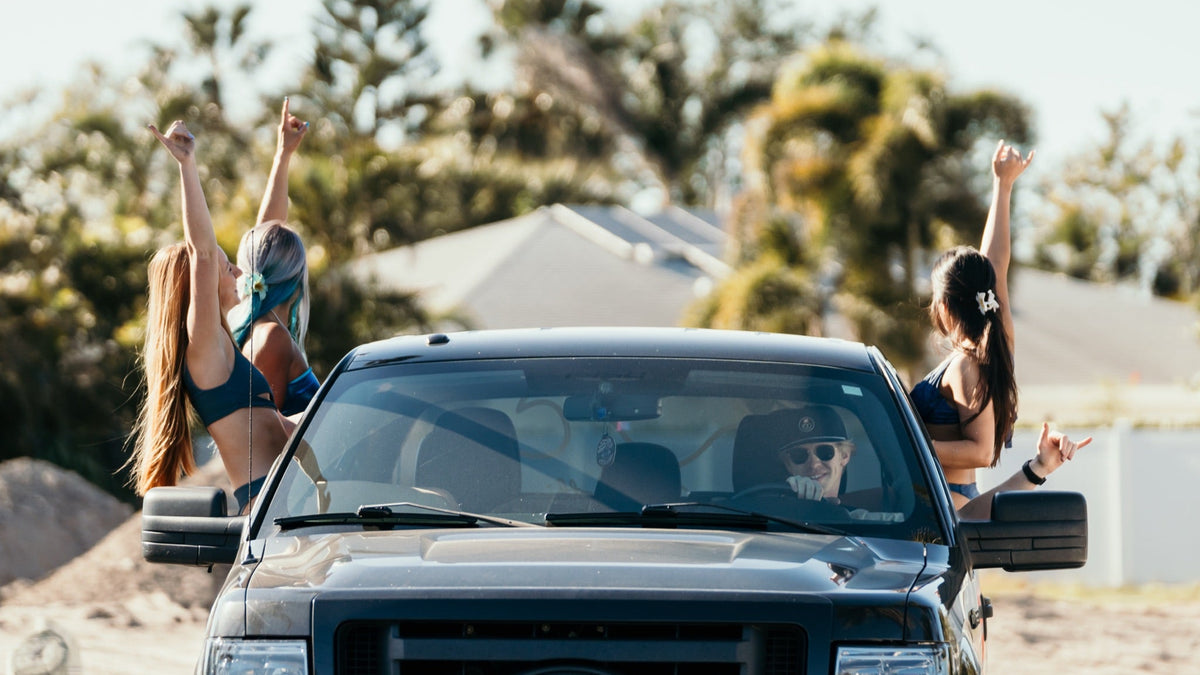 A black Ford pickup truck drives outdoors with two women leaning out of the side windows, raising their arms in a sunny, beach-like setting with palm trees and houses in the background.