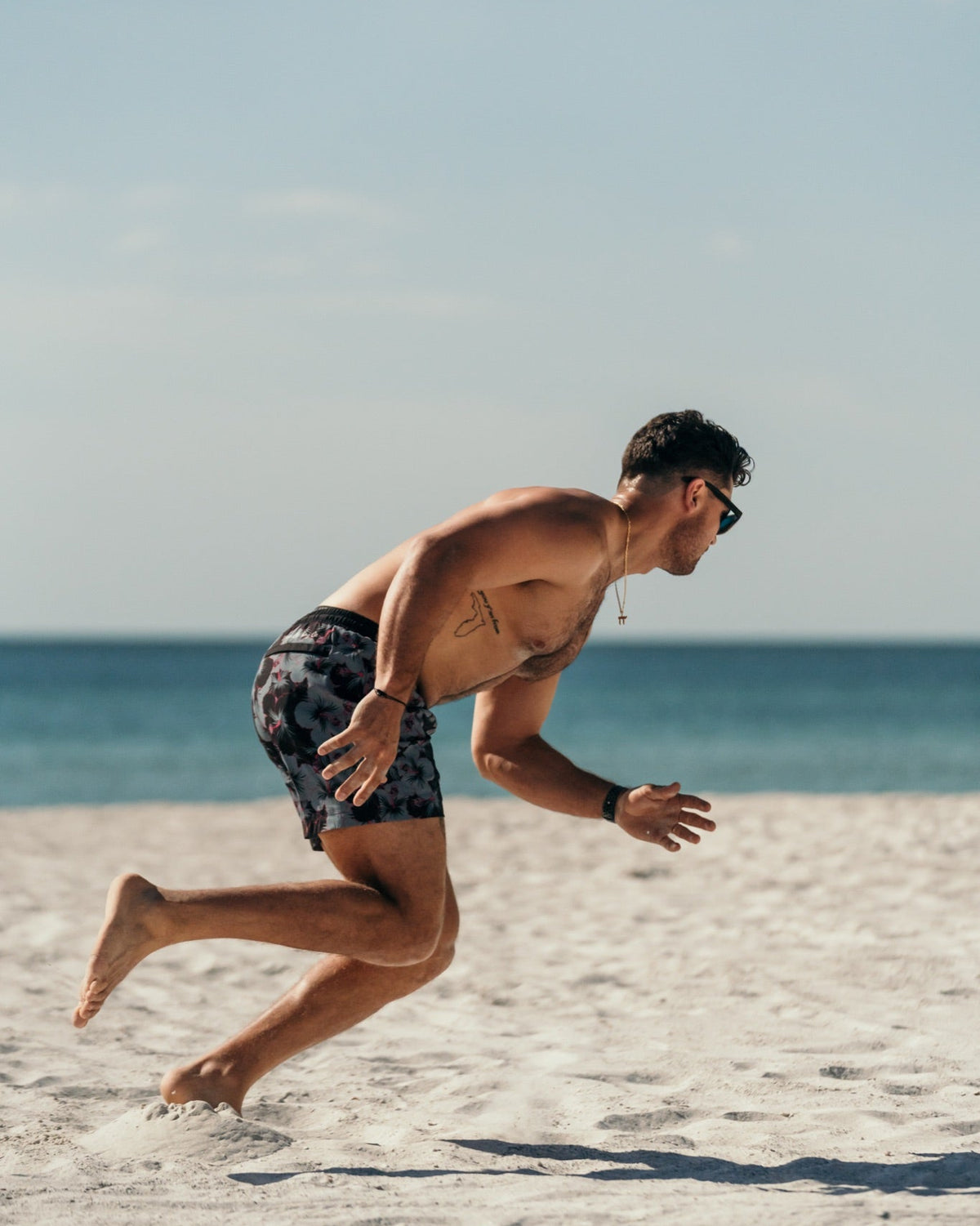 A man running on a sandy beach wearing patterned swim trunks, with the ocean and blue sky in the background.