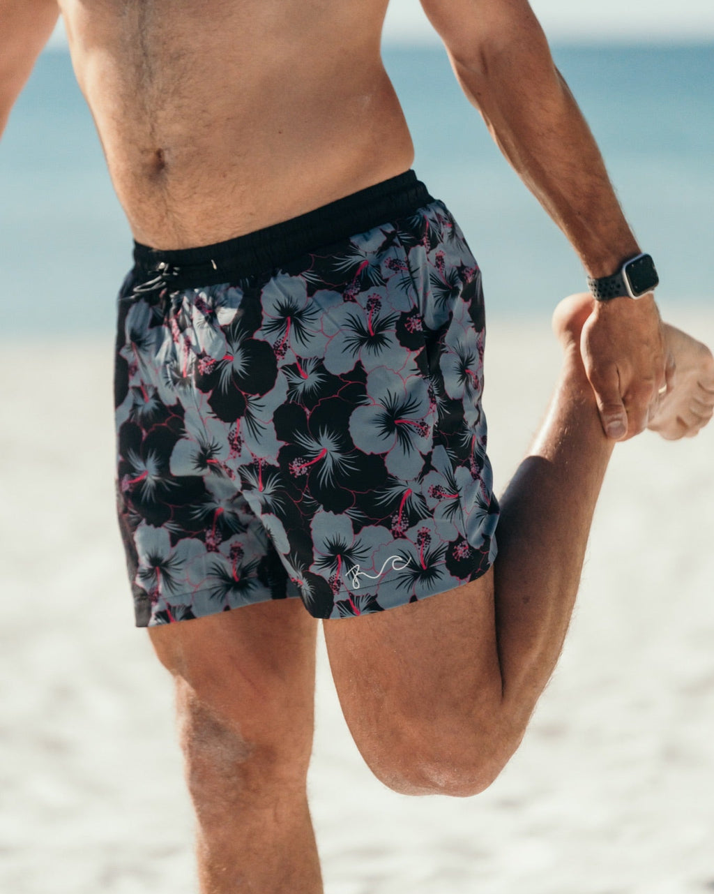 A man on a beach wearing floral swim trunks with a black waistband, stretching his leg while holding another person's hand. The swim trunks feature a dark background with light gray, pink, and black tropical floral patterns.
