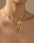 A gold four-petal flower pendant necklace worn on a model's neck, shown against a warm beige background.