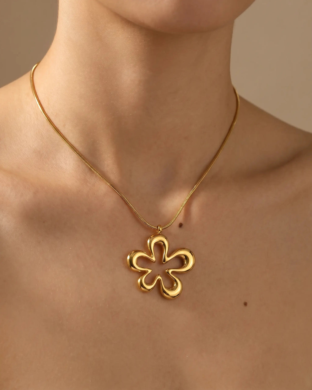 A gold four-petal flower pendant necklace worn on a model's neck, shown against a warm beige background.