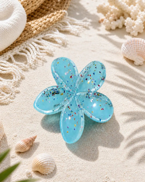 A translucent light blue flower-shaped hair clip with embedded glitter, displayed on a sandy beach with seashells, a woven hat, and ocean water in the background.
