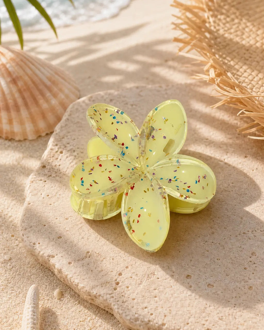 A translucent pale yellow flower-shaped hair clip with multicolored glitter specks, displayed on a stone slice over a sandy beach setting with seashells, a starfish, and ocean waves in the background.