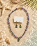 A beaded necklace and matching earrings displayed on sand with seashells and palm leaves. The necklace and earrings feature lavender purple beads, iridescent blue-green beads, and gold-tone hardware, including a turtle-shaped pendant and starfish-shaped earrings.
