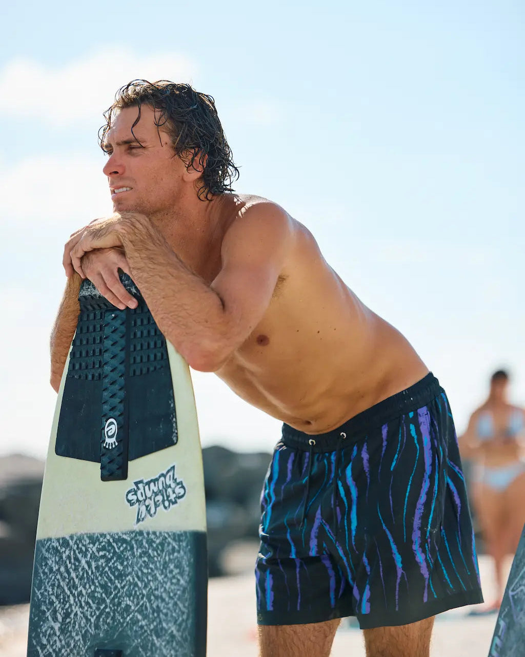 A shirtless man on a beach leaning on a surfboard, wearing dark patterned swim trunks with blue and purple wavy stripes. The surfboard has visible 'Surf Life' branding.