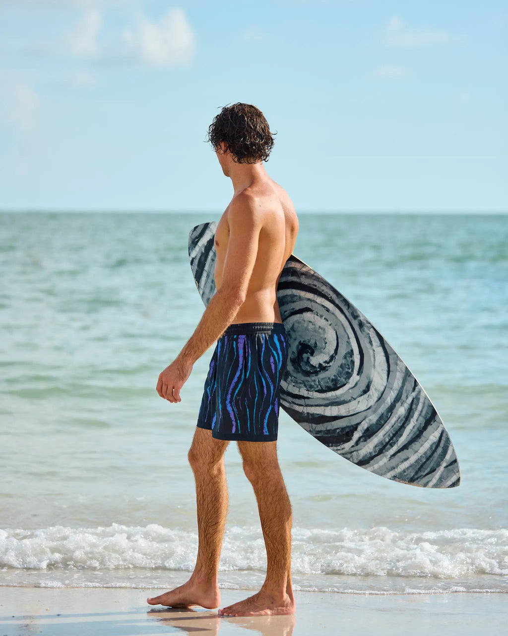 A man standing on a sandy beach at the shoreline, seen from the back, wearing blue patterned swim trunks and carrying a black and white swirled surfboard under his arm.