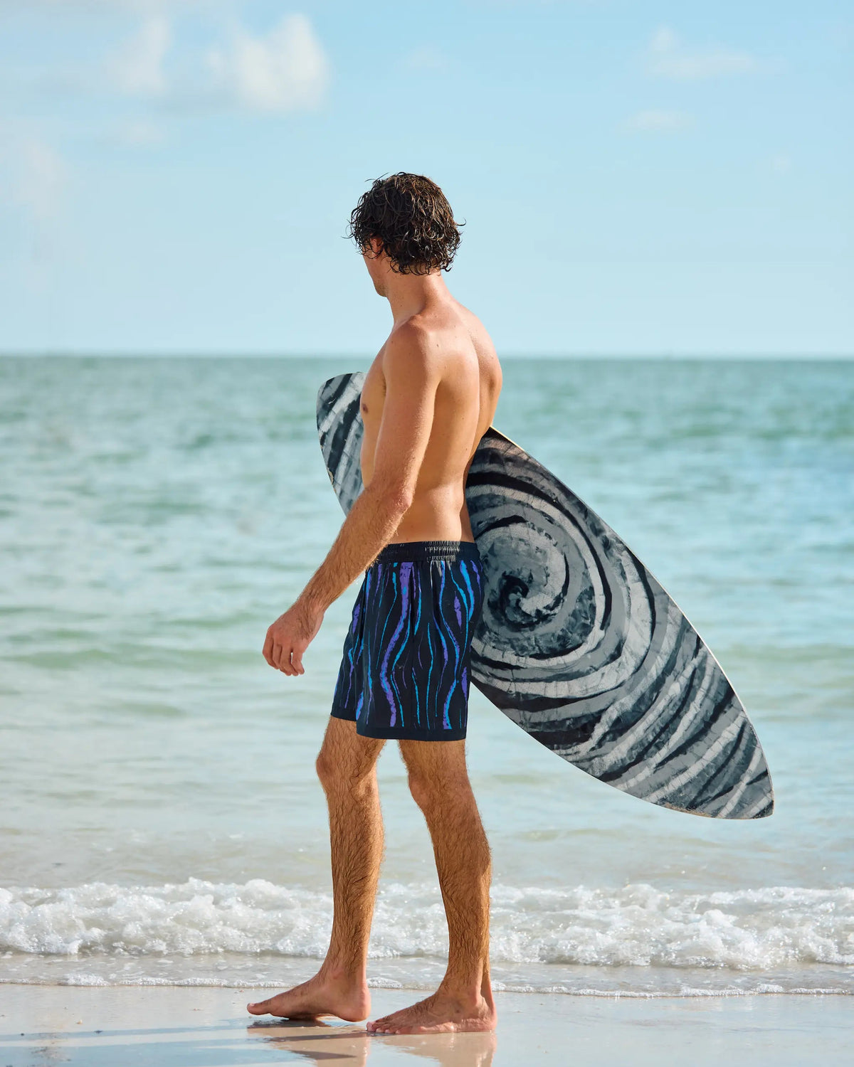 A man standing on a sandy beach at the shoreline, seen from the back, wearing blue patterned swim trunks and carrying a black and white swirled surfboard under his arm.