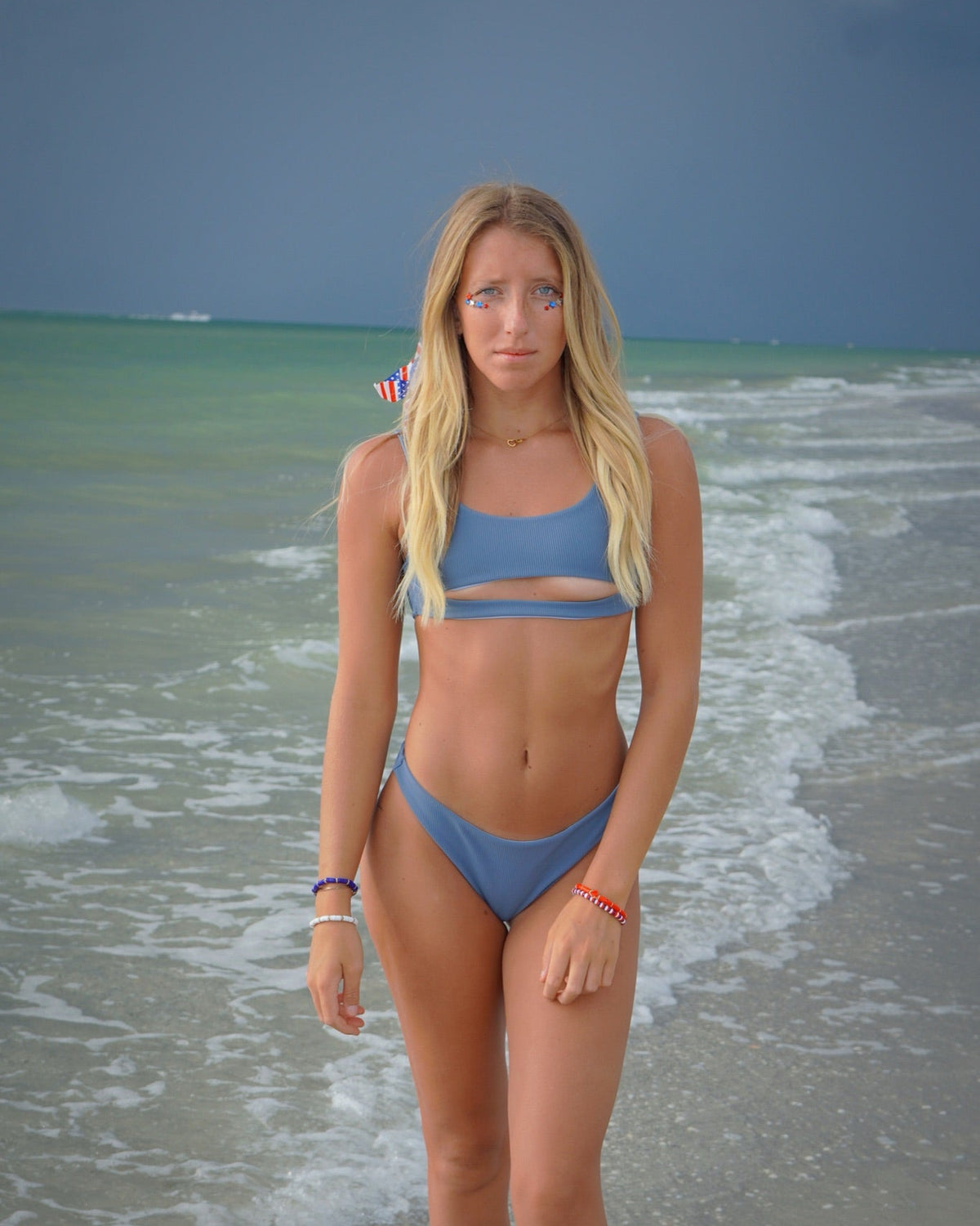 A woman with long blonde hair stands on a beach wearing a light blue two-piece bikini. The background shows ocean waves and a dark blue sky.