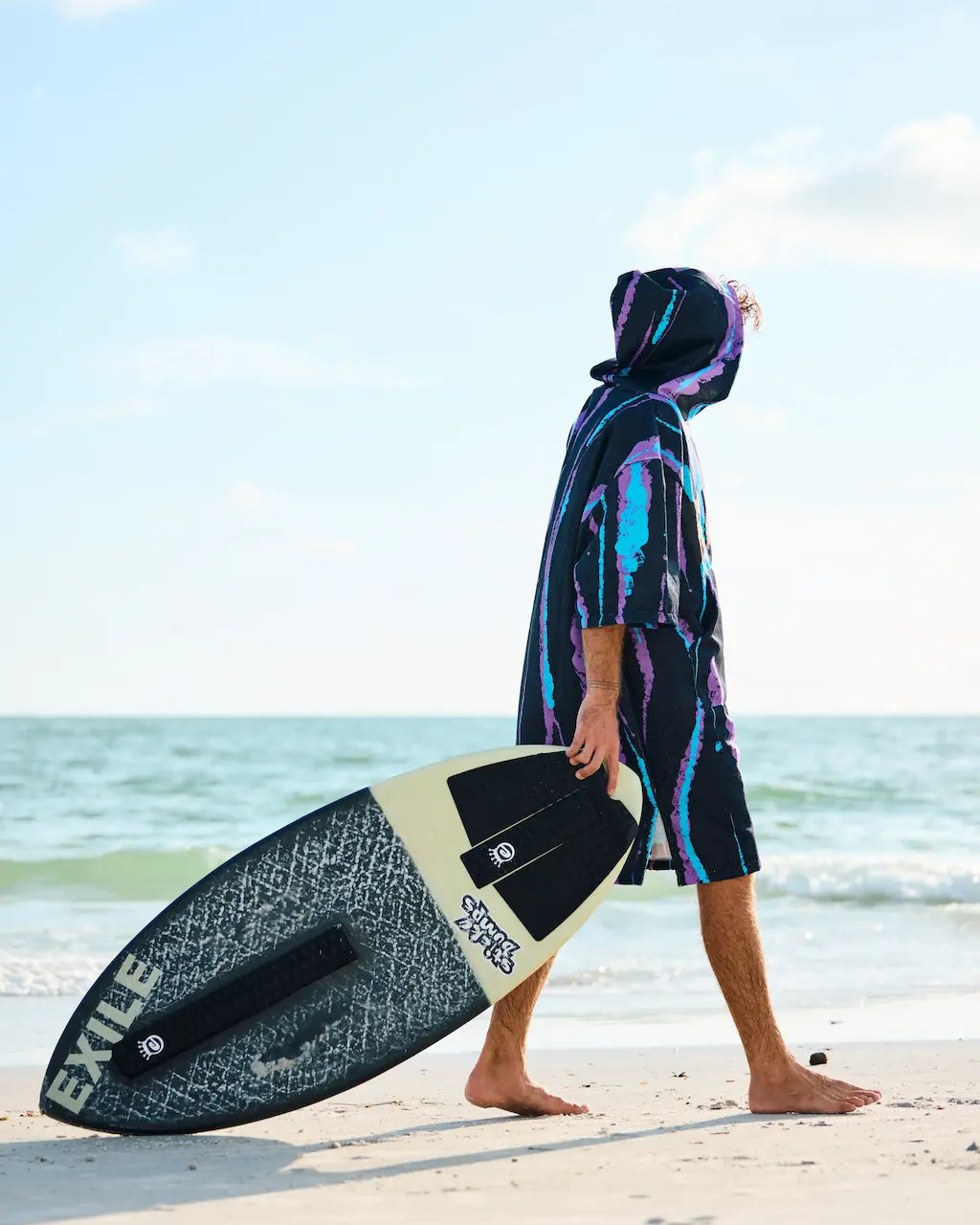 A person walks along a sandy beach carrying a surfboard, wearing a dark hooded poncho-style cover with bright blue, purple, and pink tie-dye-like streaks. The ocean and sky are visible in the background.