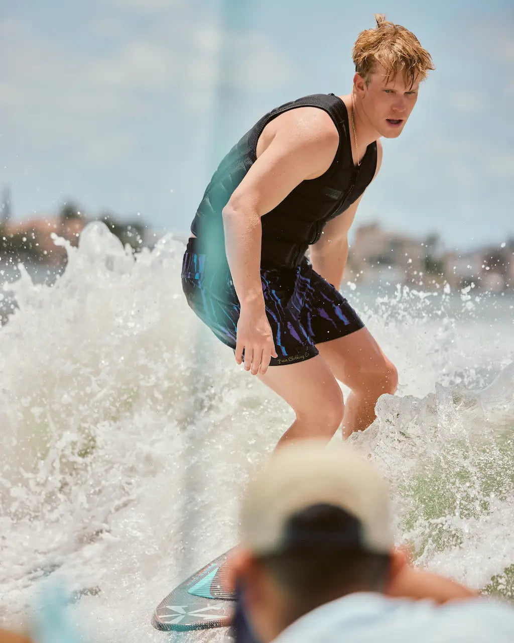 A man surfing on a wave in an ocean, wearing a black sleeveless top and dark patterned shorts, with water splashing around him and a blurred person in the foreground.