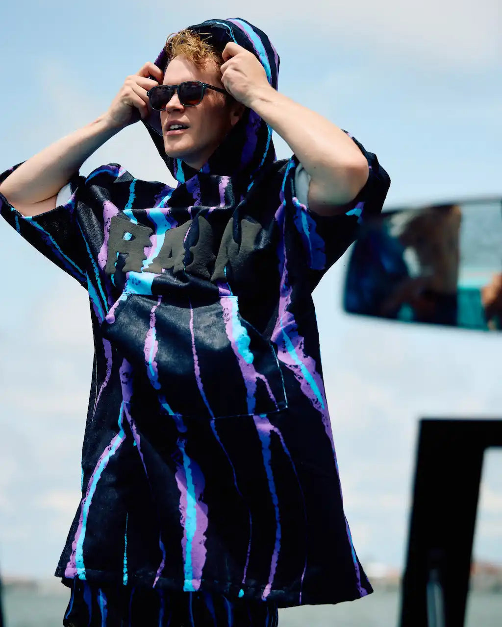 A man wearing a black oversized hoodie with purple and blue tie-dye streaks, matching black shorts, and sunglasses, standing outdoors with his hands on his hood.