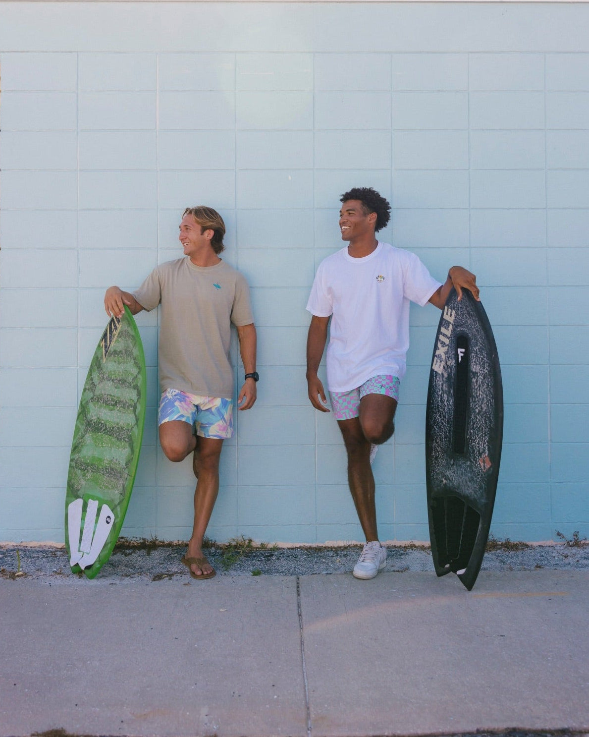 Two men standing against a light blue brick wall outdoors, each holding a surfboard. One man wears a beige t-shirt and patterned shorts, holding a green surfboard.