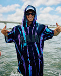 A man wearing a black hooded poncho with purple and blue vertical streaks stands on a boat with water and a bridge in the background, giving two thumbs up.