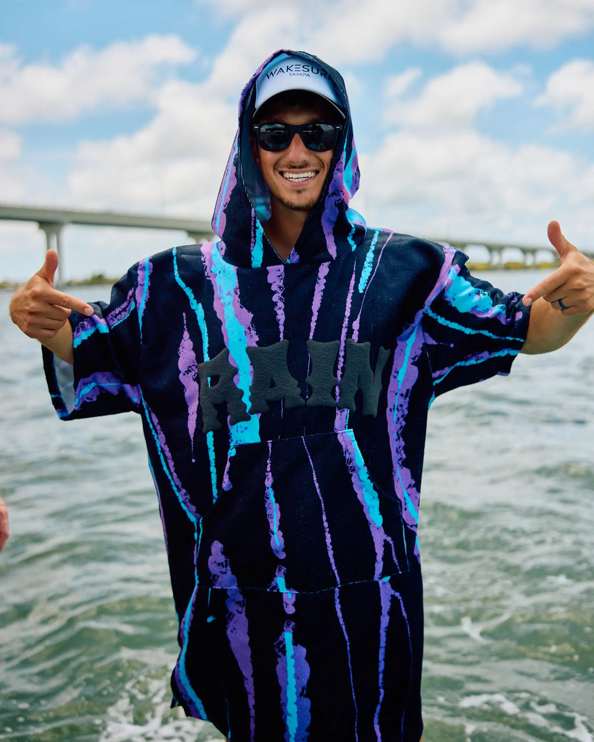 A man wearing a black hooded poncho with purple and blue vertical streaks stands on a boat with water and a bridge in the background, giving two thumbs up.