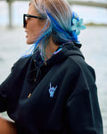 A woman with blue and blonde hair wearing a black hoodie with a small light blue hand graphic on the chest, black sunglasses, and a blue flower hair accessory.