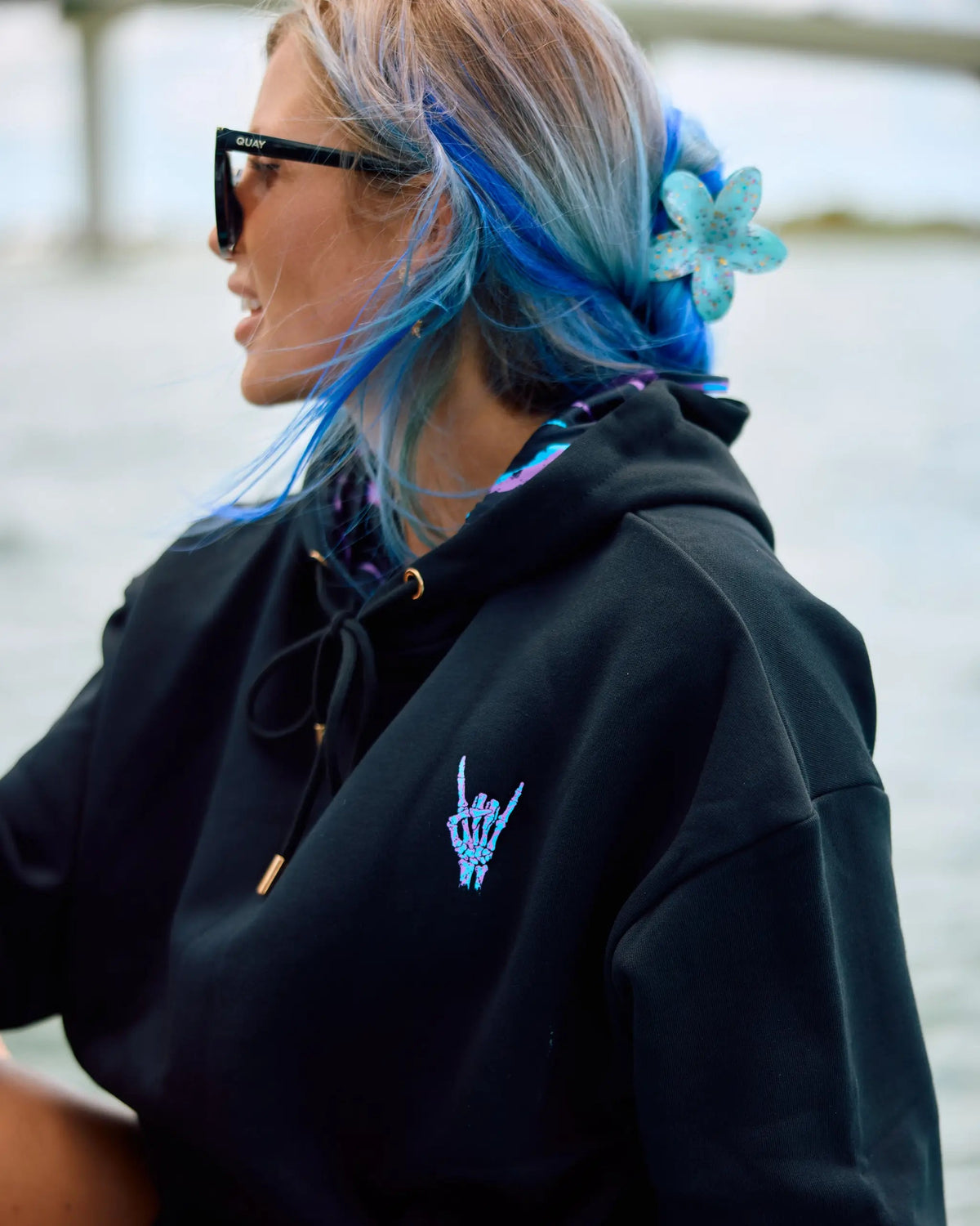 A woman with blue and blonde hair wearing a black hoodie with a small light blue hand graphic on the chest, black sunglasses, and a blue flower hair accessory.