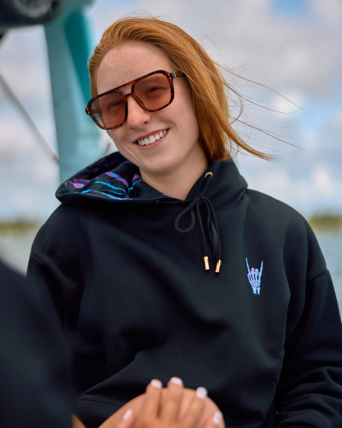 A smiling woman with red hair wearing oversized brown sunglasses and a black hoodie with a small blue graphic on the chest, photographed outdoors with a cloudy sky and blue structure in the background.