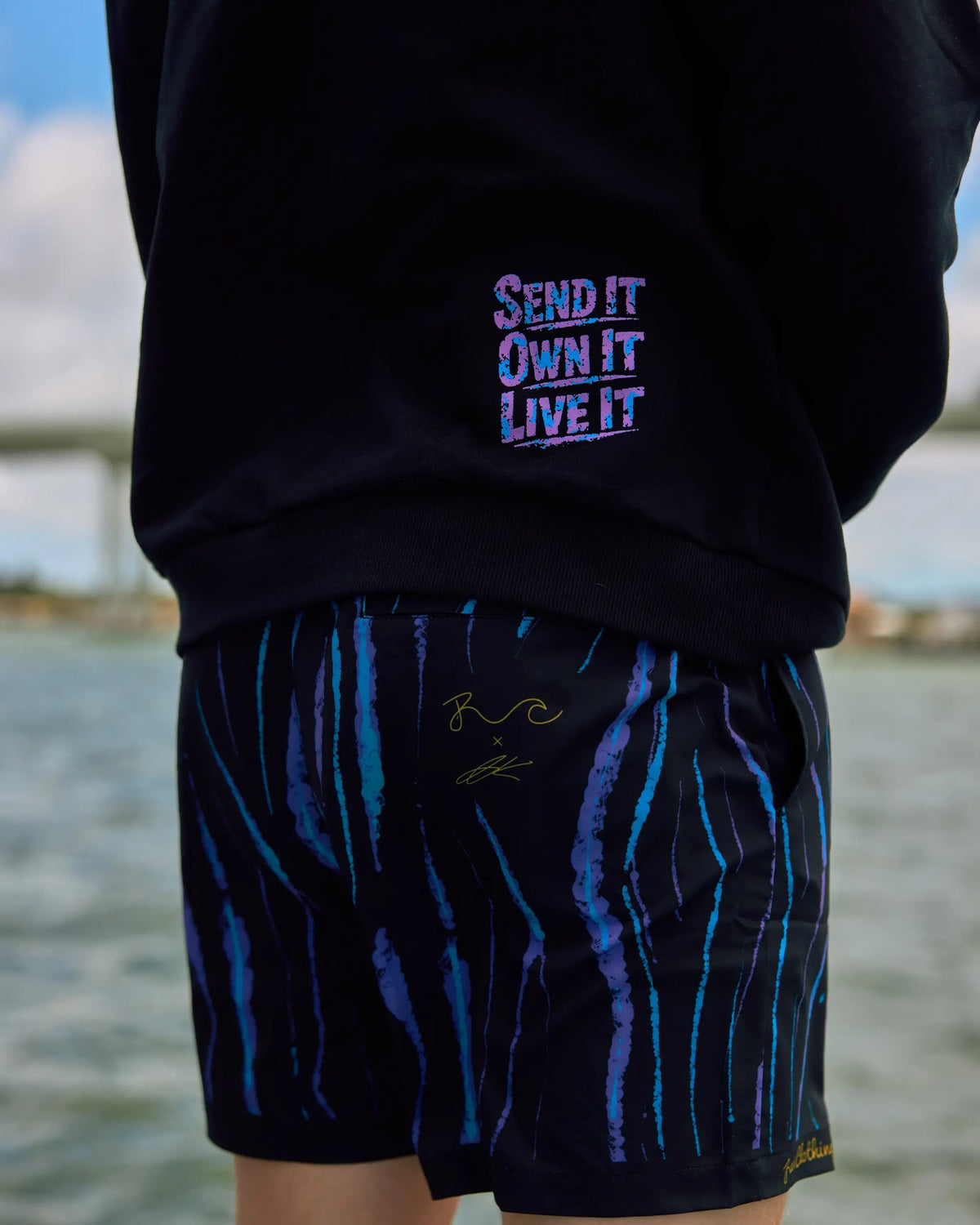 A person outdoors wearing black shorts with vertical blue and purple tiger-stripe patterns and a black sweatshirt with purple text reading 'SEND IT OWN IT LIVE IT' on the back.