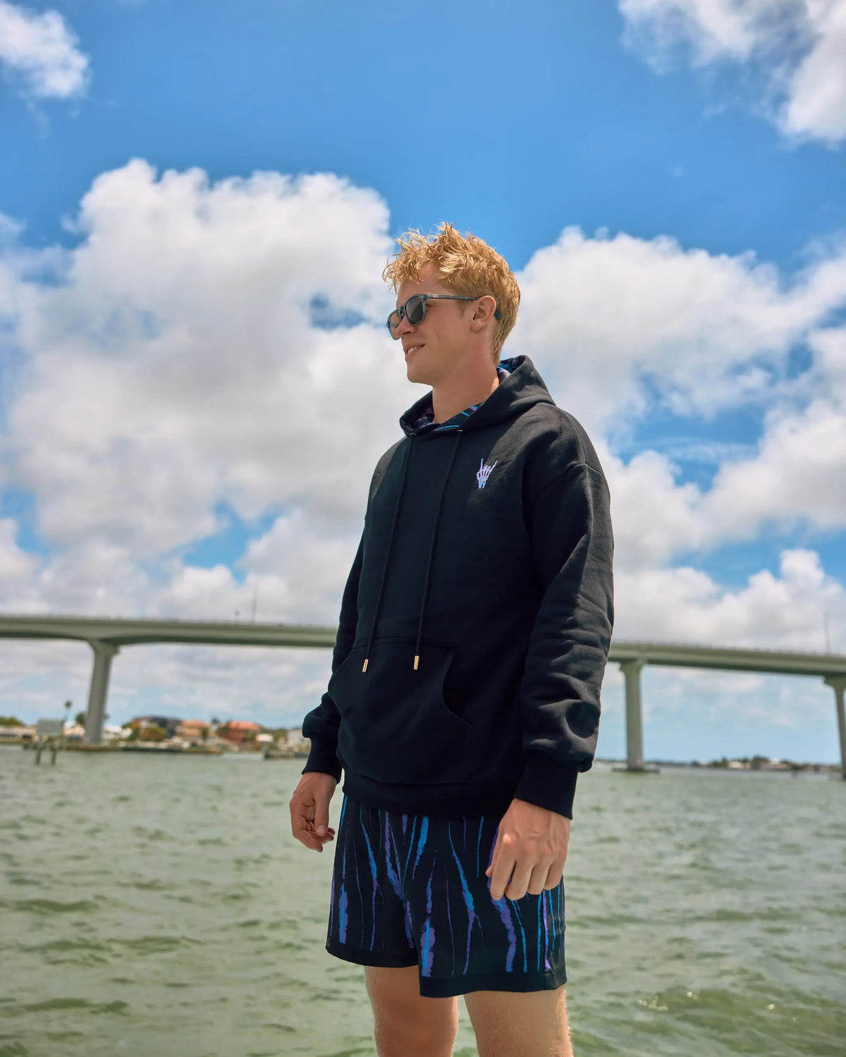 A man wearing a black pullover hoodie and blue patterned swim trunks stands outdoors near the water under a bright blue sky with white clouds.