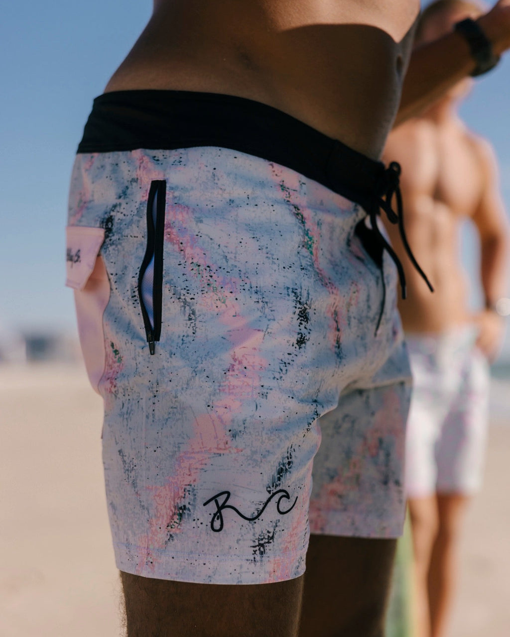 Close-up of a person wearing swim trunks at the beach. The trunks are light pink, white, and blue marbled with a black waistband, black drawstring, and a black script logo near the bottom hem.