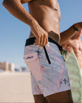 A man on a beach wearing Billabong board shorts with a white base, pastel pink and blue watercolor-like pattern, and a black waistband.