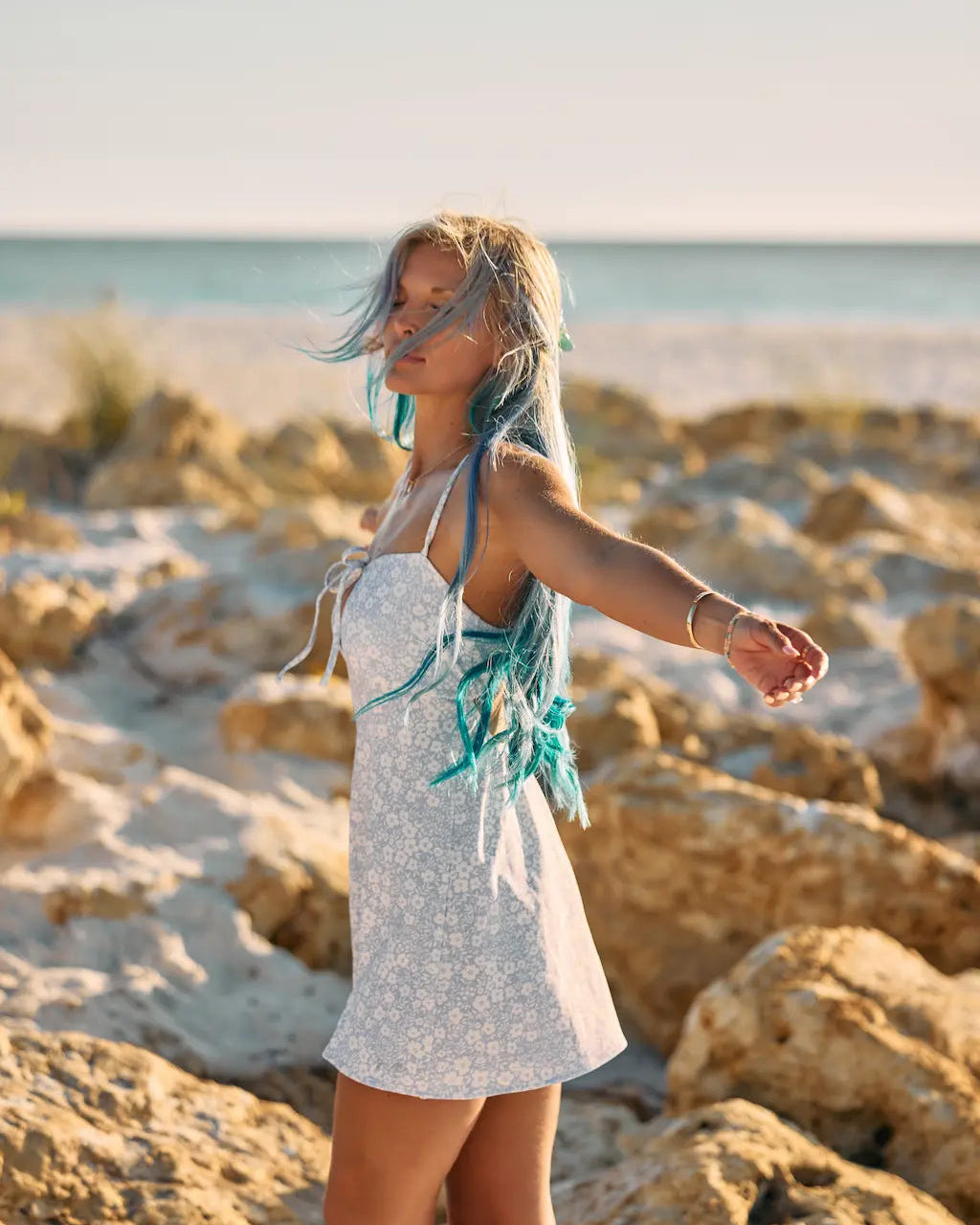 A woman stands on a rocky beach wearing a white sleeveless dress with turquoise fringe details. The background features sandy rocks, a calm sea, and a soft, warm sky.