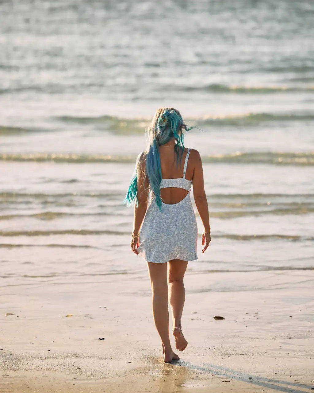 A woman with long blue hair walks barefoot on a sandy beach away from the camera, wearing a white patterned mini dress with a cutout back, with the ocean in the background.