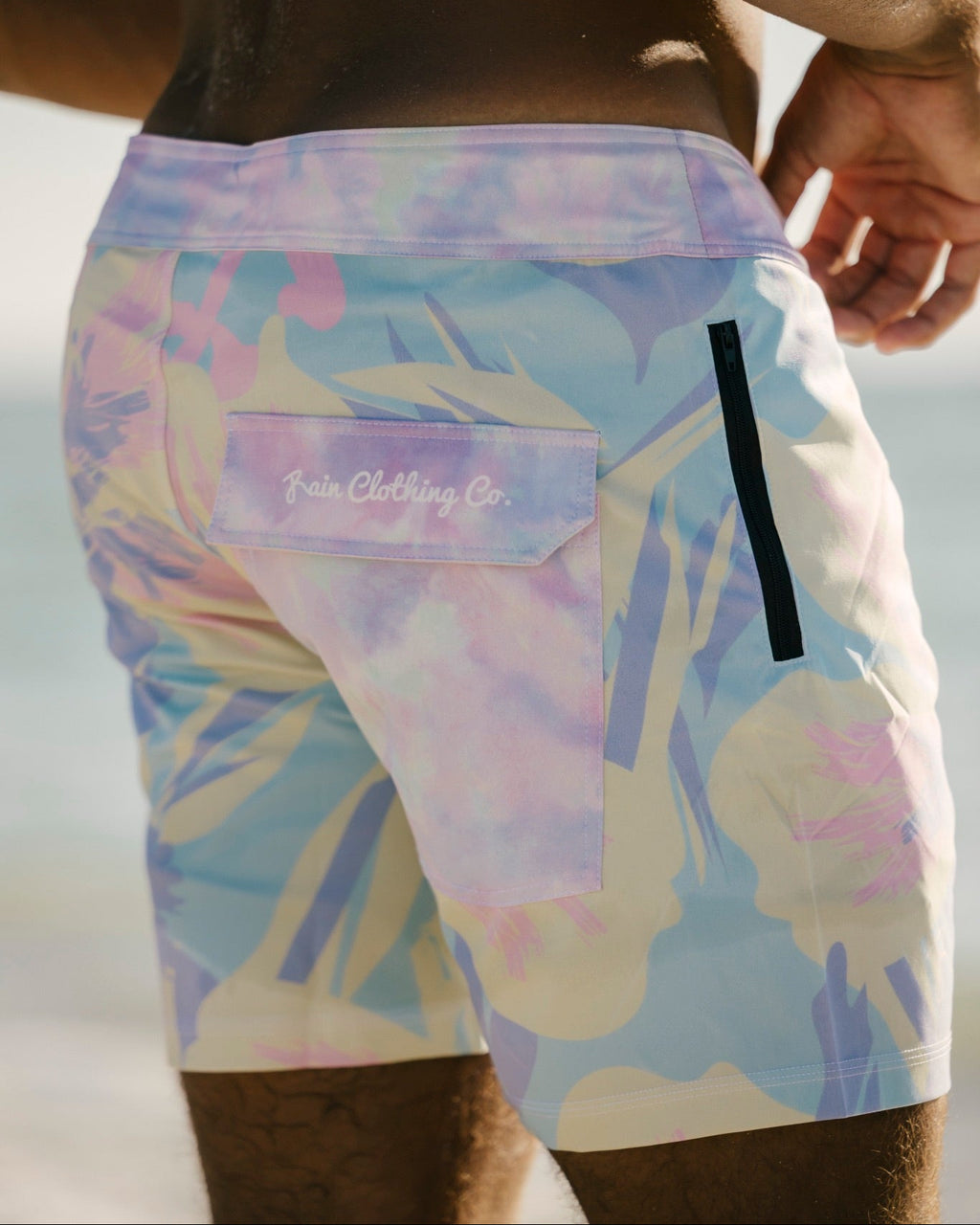 Close-up back view of a person wearing swim trunks with a pastel tie-dye pattern in lavender, blue, pink, and cream. The shorts have a side pocket with the 'Fair Clothing Co.