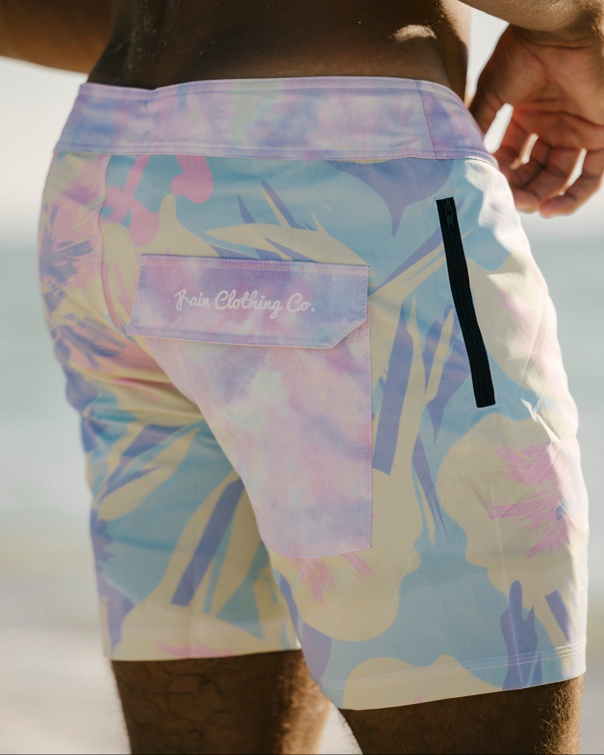 Close-up back view of a person wearing swim trunks with a pastel tie-dye pattern in lavender, blue, pink, and cream. The shorts have a side pocket with the 'Fair Clothing Co.