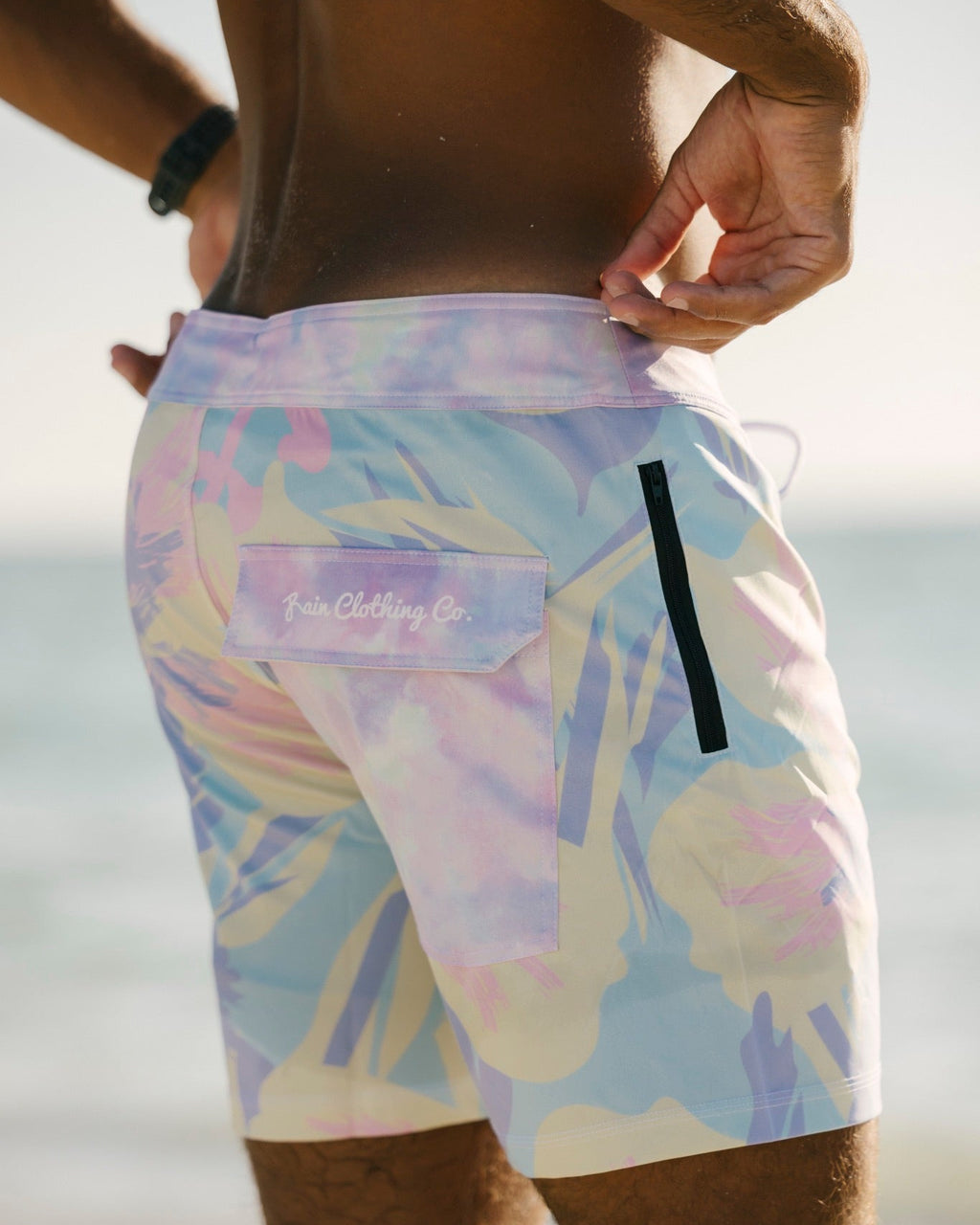 A close-up back view of a person wearing pastel abstract print board shorts with a visible 'Tune Clothing Co.' logo patch on the back pocket, photographed outdoors near water.