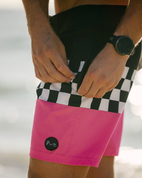 Close-up of a person wearing bright pink board shorts with a black-and-white checkered waistband and a small circular logo patch near the hem, photographed outdoors with a blurred light background.