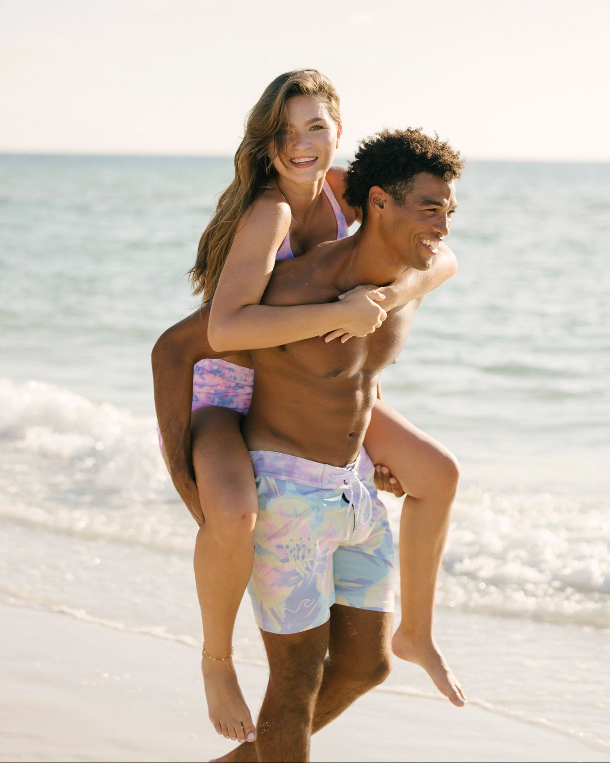 A smiling couple on a beach, with the man carrying the woman on his back. The woman wears a light purple bikini top and patterned pastel swim trunks, and the man wears light blue and white patterned swim trunks.