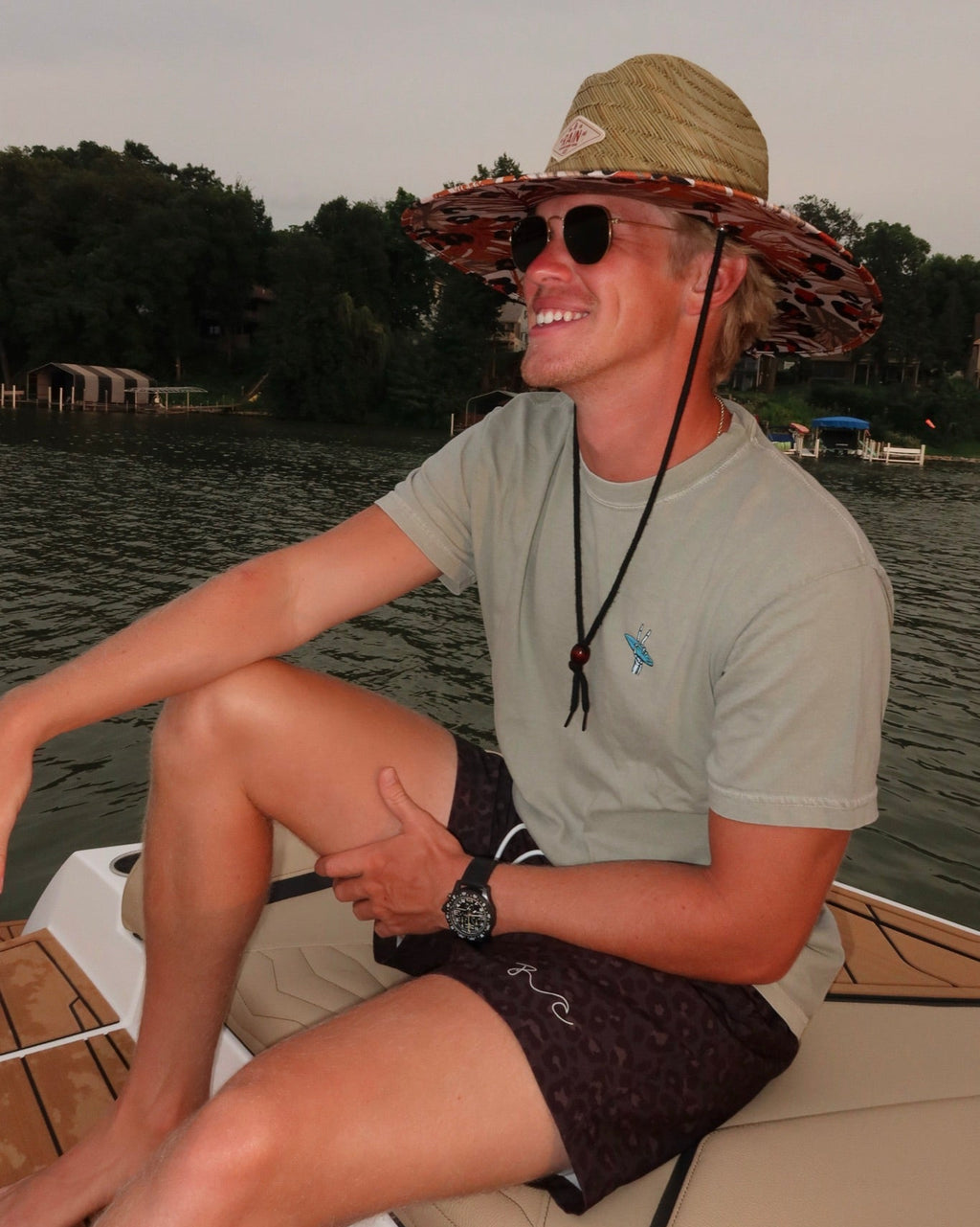A smiling man sitting on a boat wearing a wide-brimmed camouflage hat, sunglasses, a light gray t-shirt, and dark patterned shorts, with water and trees in the background.