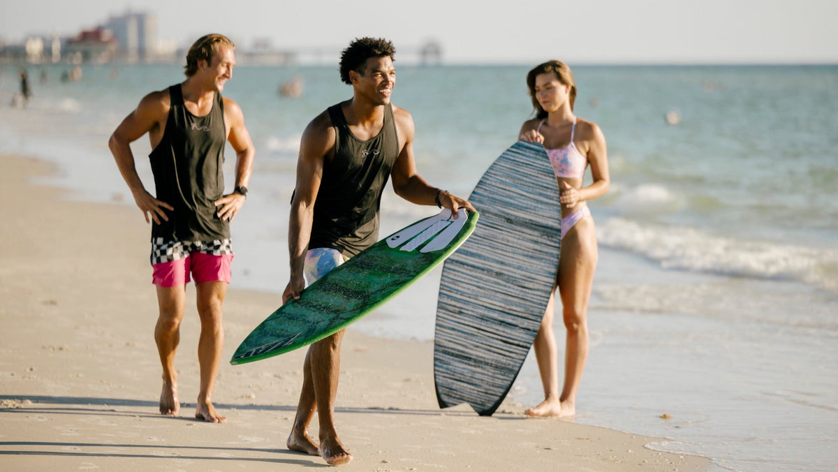 Three people walking on a sandy beach near the ocean, each carrying a surfboard. The person on the left wears a black tank top and pink shorts, the person in the middle wears a black tank top and light blue swim trunks while holding a green surfboard, and the person on the right wears a light pink bikini while holding a gray striped surfboard.